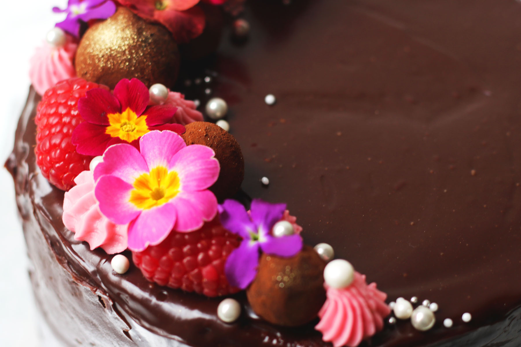 Close up of raspberries and edible flowers on top of a chocolate ganache covered cake