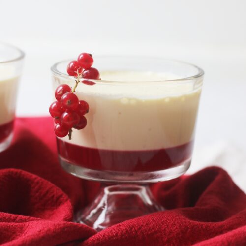 A pudding glass filled with a redcurrant jelly topped with an orange posset. Redcurrants garnish the dessert.