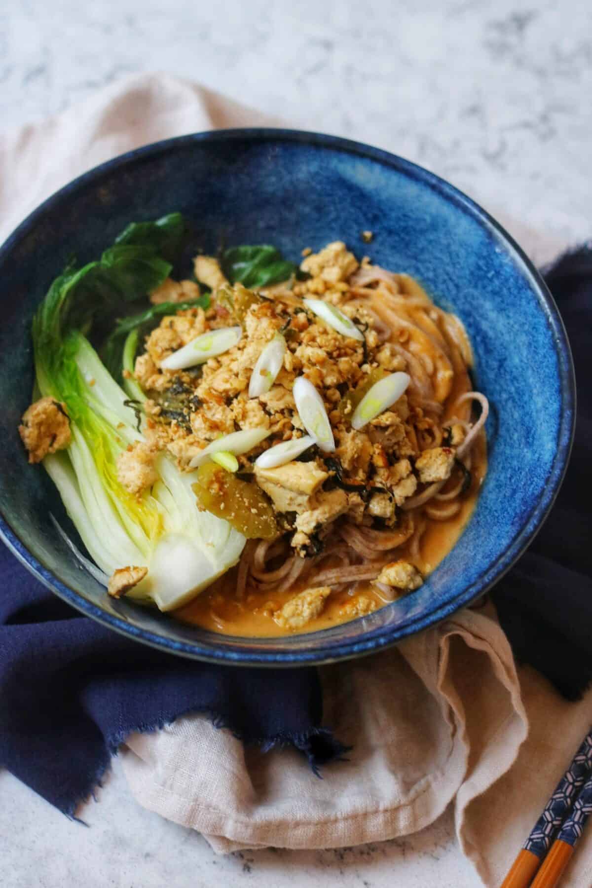 A bowl of vegan dan dan noodles and chopsticks on a blue and cream napkin