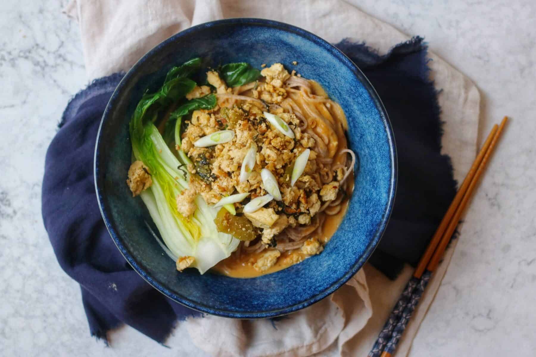 A bowl of vegan dan dan noodles and chopsticks on a blue and cream napkin