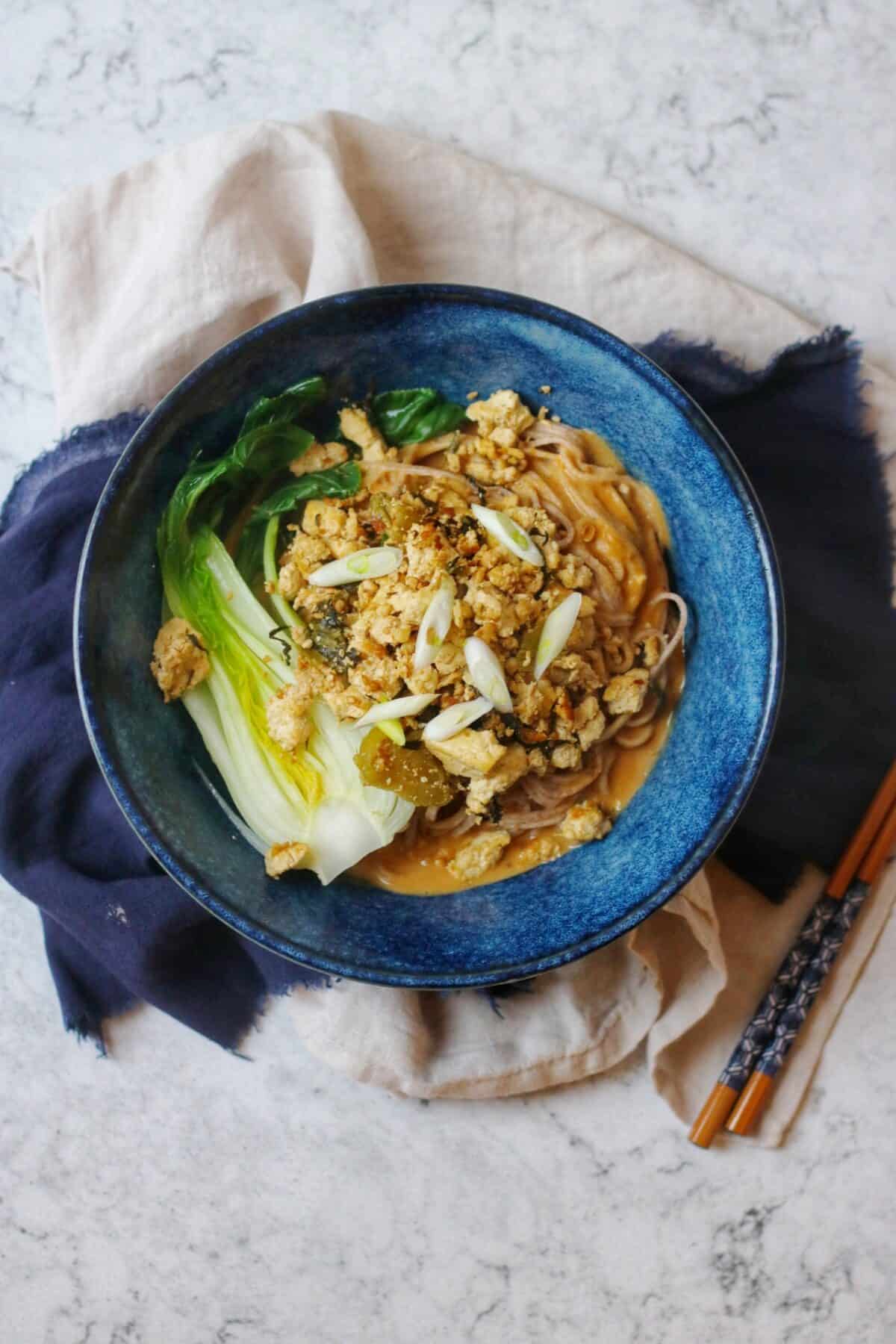 A bowl of vegan dan dan noodles and chopsticks on a blue and cream napkin