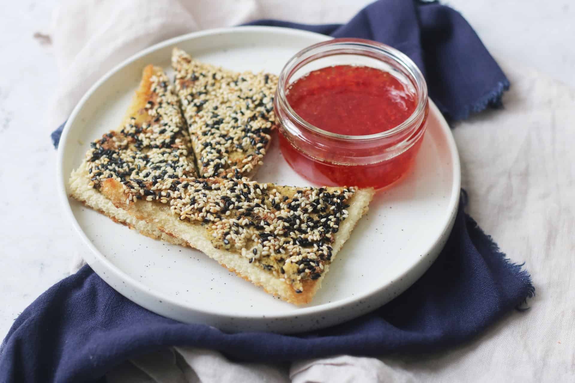 A plate with 3 triangles of vegan sesame prawn toasts and a small pot of sweet chilli sauce