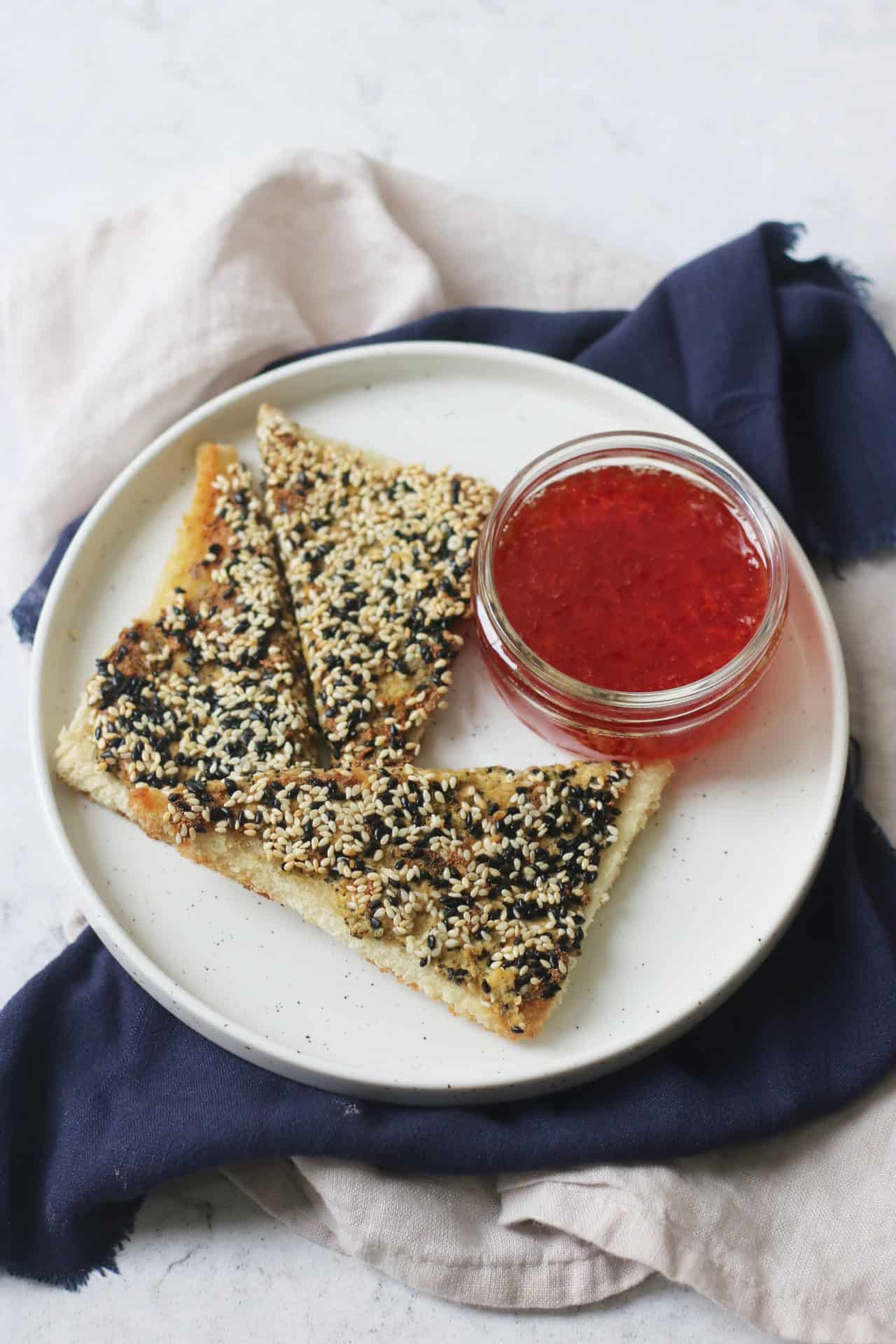 A plate with 3 triangles of vegan sesame prawn toasts and a small pot of sweet chilli sauce