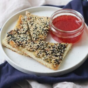 A plate with 3 triangles of vegan sesame prawn toasts and a small pot of sweet chilli sauce