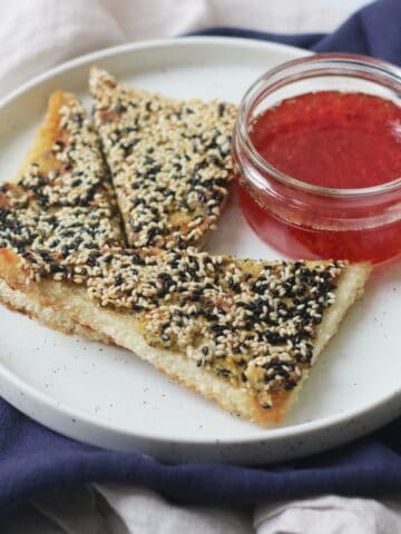 A plate with 3 triangles of vegan sesame prawn toasts and a small pot of sweet chilli sauce