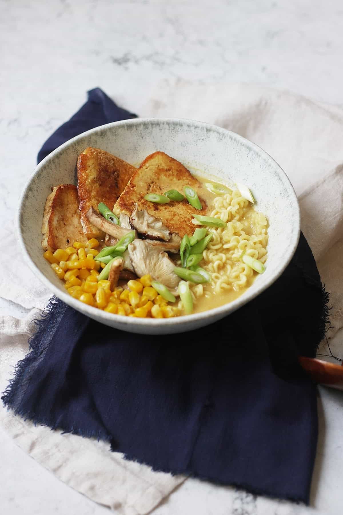 A bowl of Vegan Tonkotsu Ramen topped with smoked tofu, shitake mushrooms, sweetcorn and spring onions served in a blue bowl with soup spoon and chopsticks