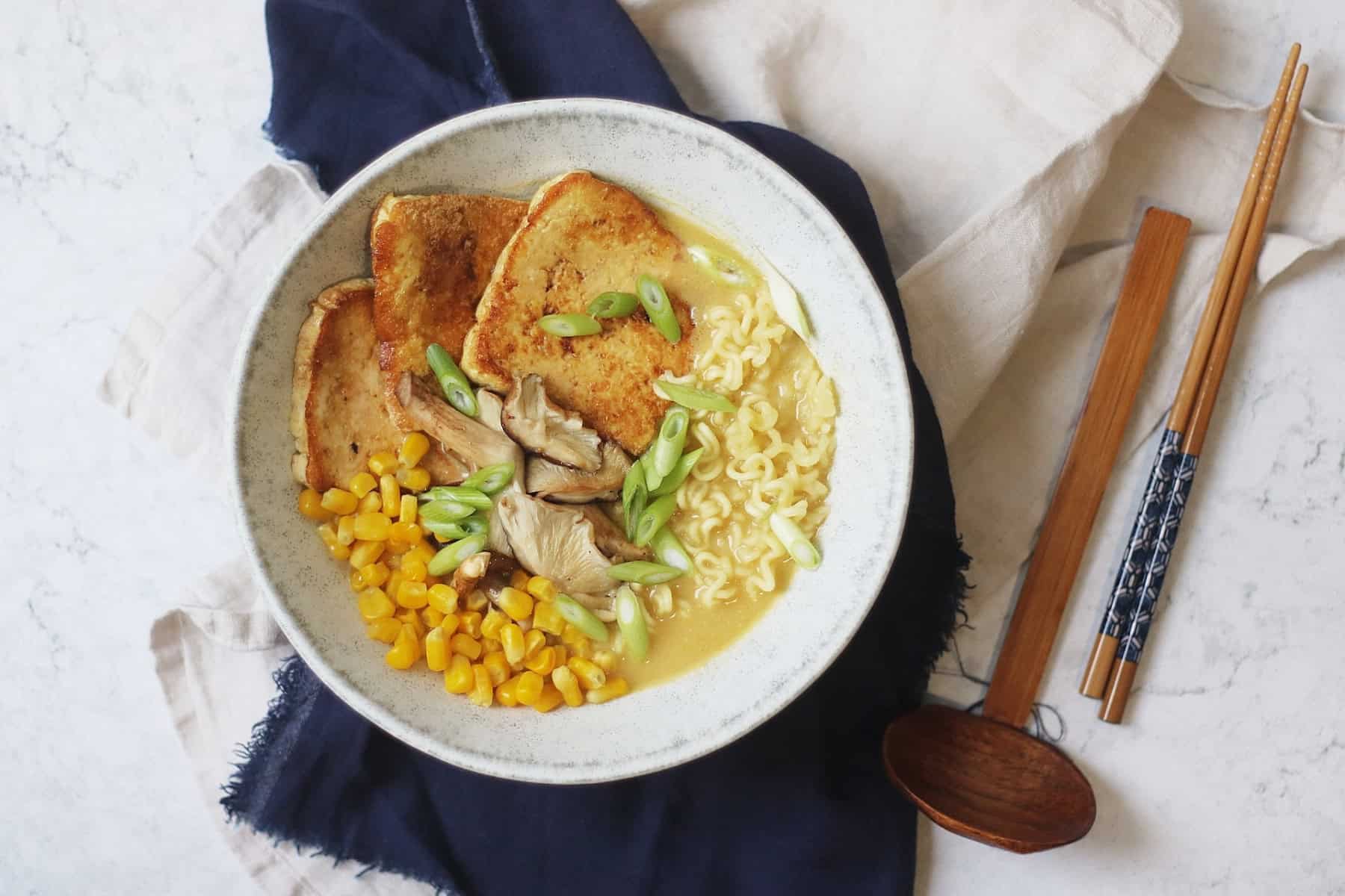 A bowl of Vegan Tonkotsu Ramen topped with smoked tofu, shitake mushrooms, sweetcorn and spring onions served in a blue bowl with soup spoon and chopsticks