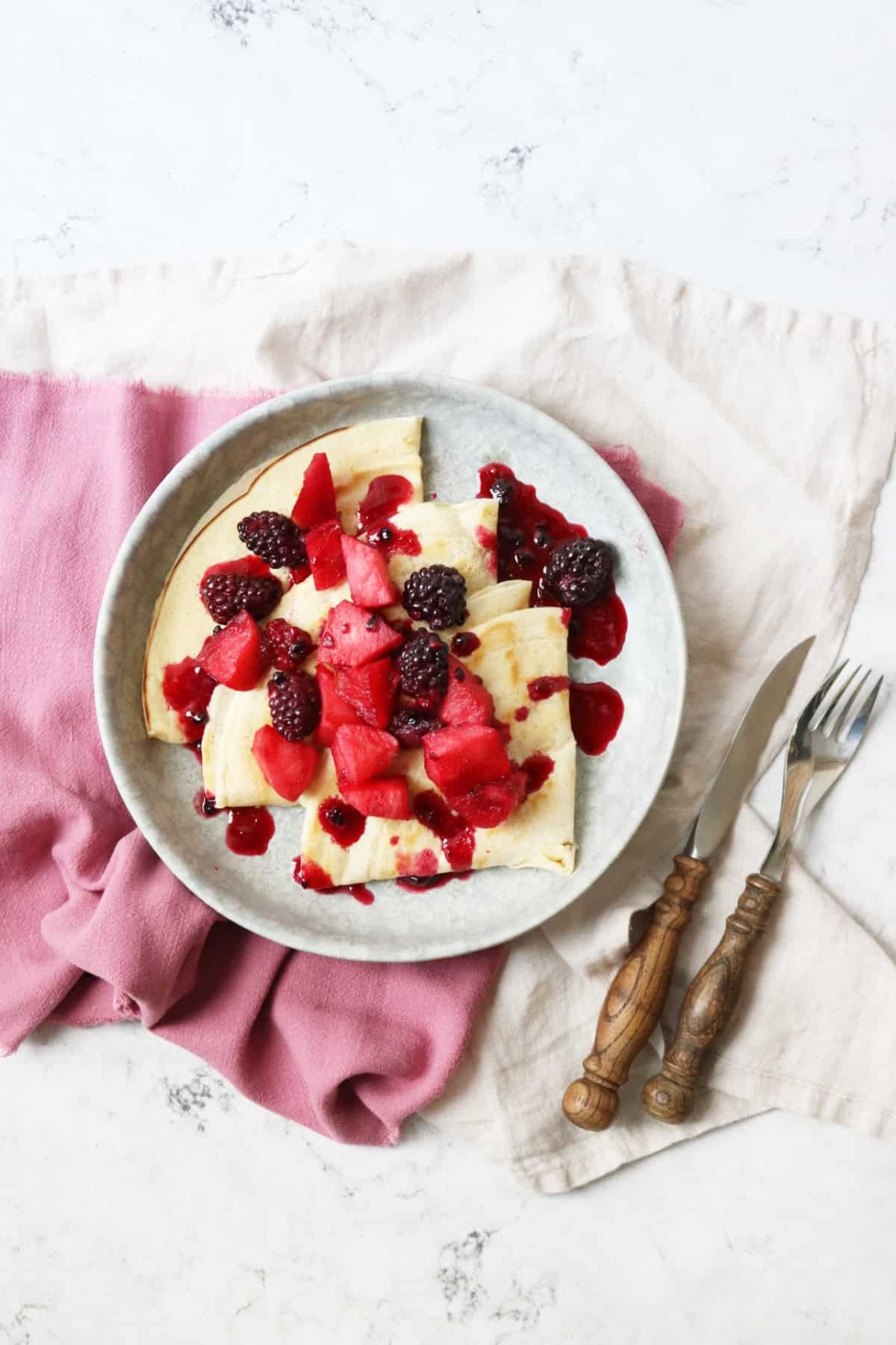 A plate of Vegan Crêpes with stewed apples and blackberries spooned over the top with a knife and fork on the side