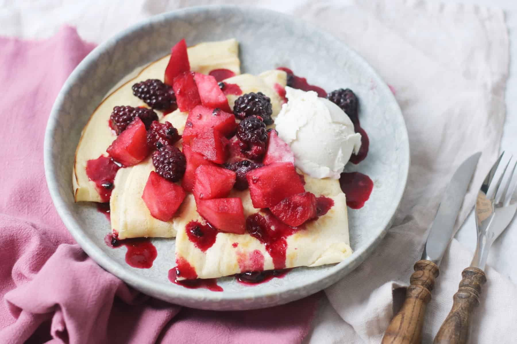 A plate of Vegan Crêpes with stewed apples and blackberries spooned over the top alongside a scoop of vegan vanilla ice cream and a knife and fork on the side