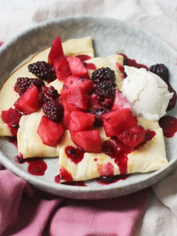 A plate of Vegan Crêpes with stewed apples and blackberries spooned over the top alongside a scoop of vegan vanilla ice cream