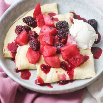 A plate of Vegan Crêpes with stewed apples and blackberries spooned over the top alongside a scoop of vegan vanilla ice cream