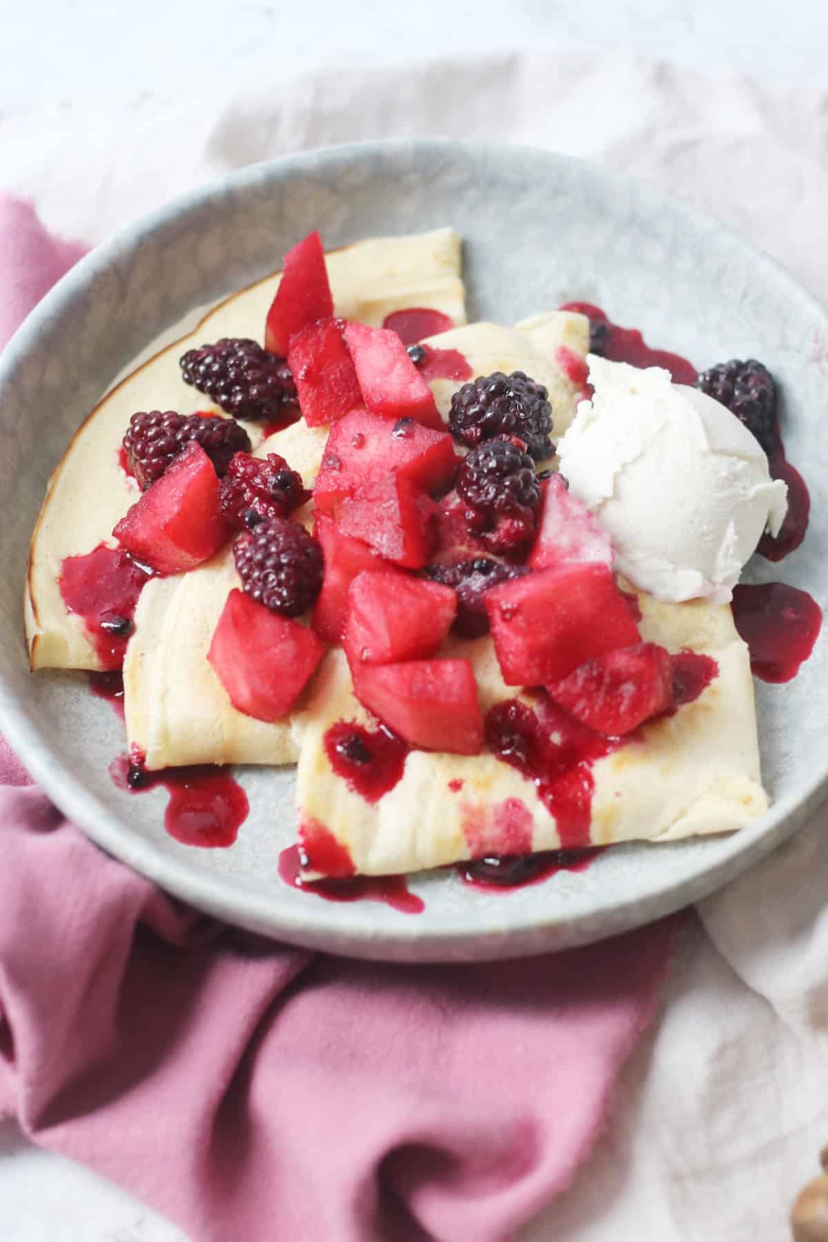 A plate of Vegan Crêpes with stewed apples and blackberries spooned over the top alongside a scoop of vegan vanilla ice cream