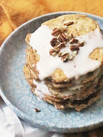 A stack of 6 vegan carrot cake pancakes topped with vegan cream cheese frosting and chopped pecan nuts.