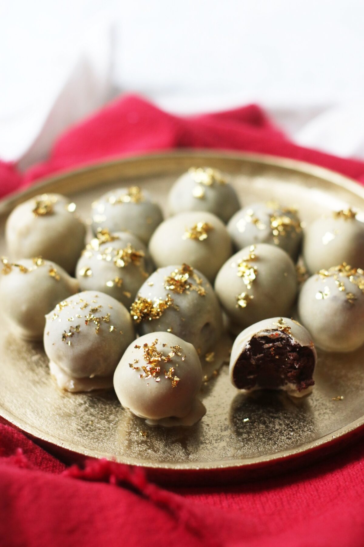 Vegan white chocolate and champagne truffles topped with gold leaf on a gold plate and red napkin. One truffle has a bite taken out to show the dark chocolate centre.