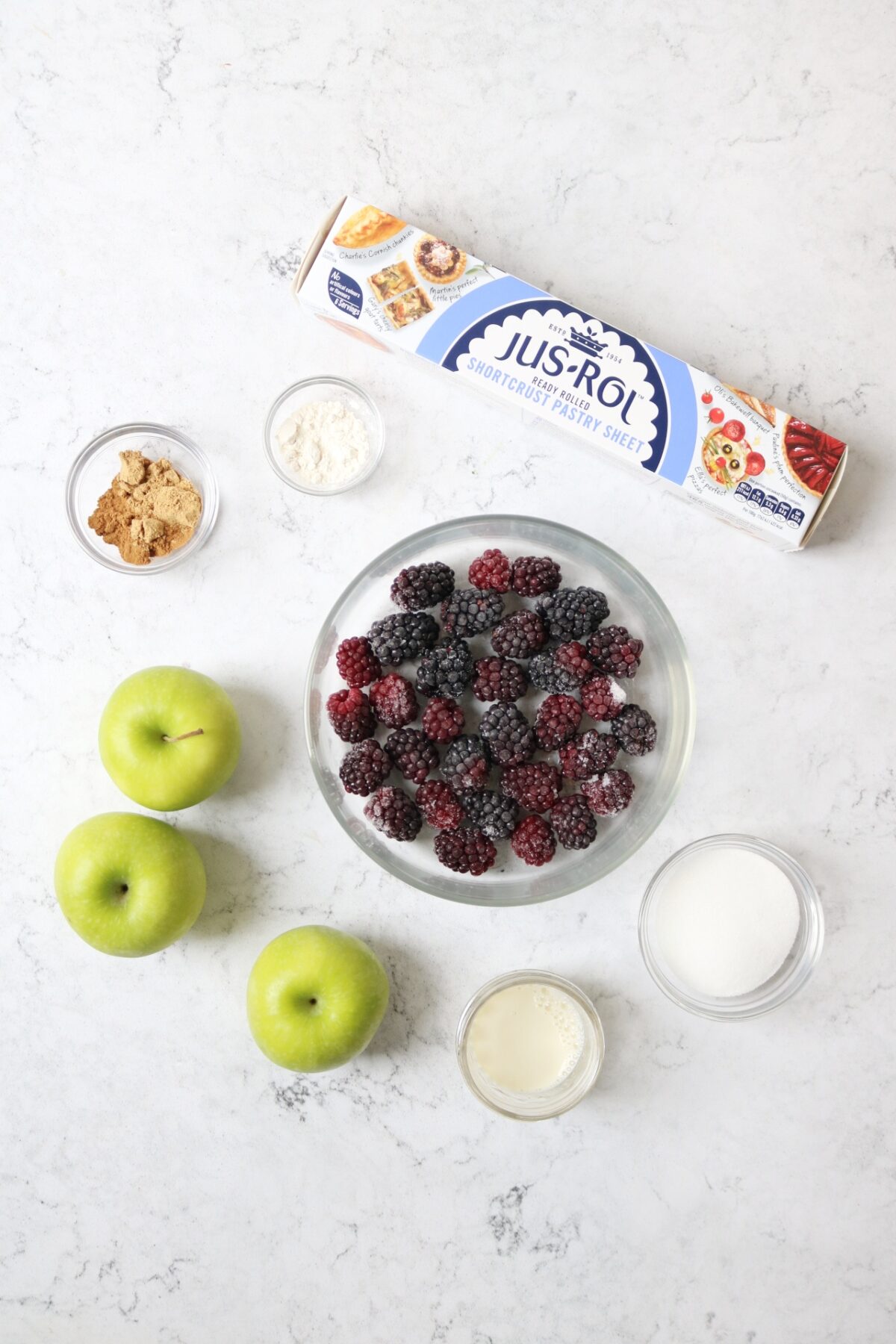 Shortcrust pastry, ground cinnamon and ginger, cornflour, plant milk, caster sugar, 3 apples, a packet of shortcrust pastry and a bowl of blackberries laid out on a white marble counter top