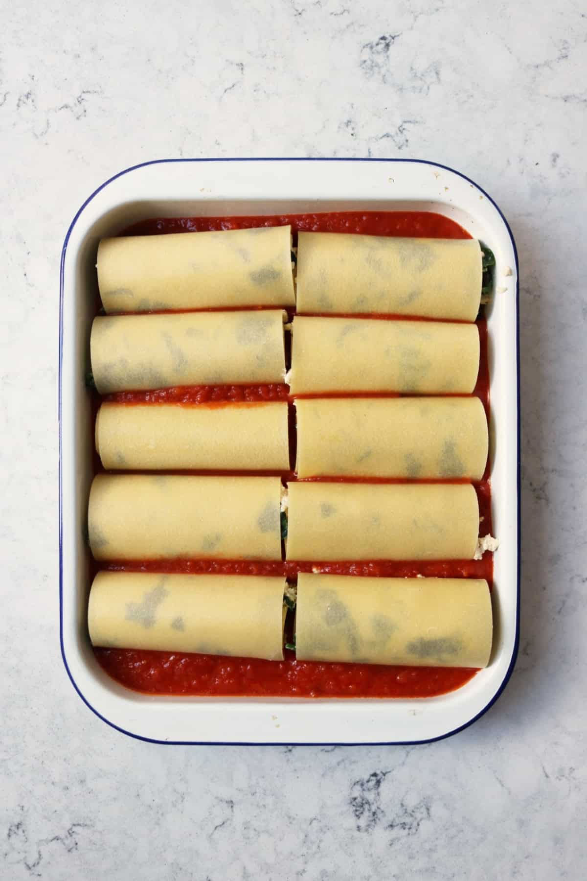 A blue and white enamel oven tin filled with tomato sauce and rolled up lasagne sheets stuffed with spinach and vegan ricotta