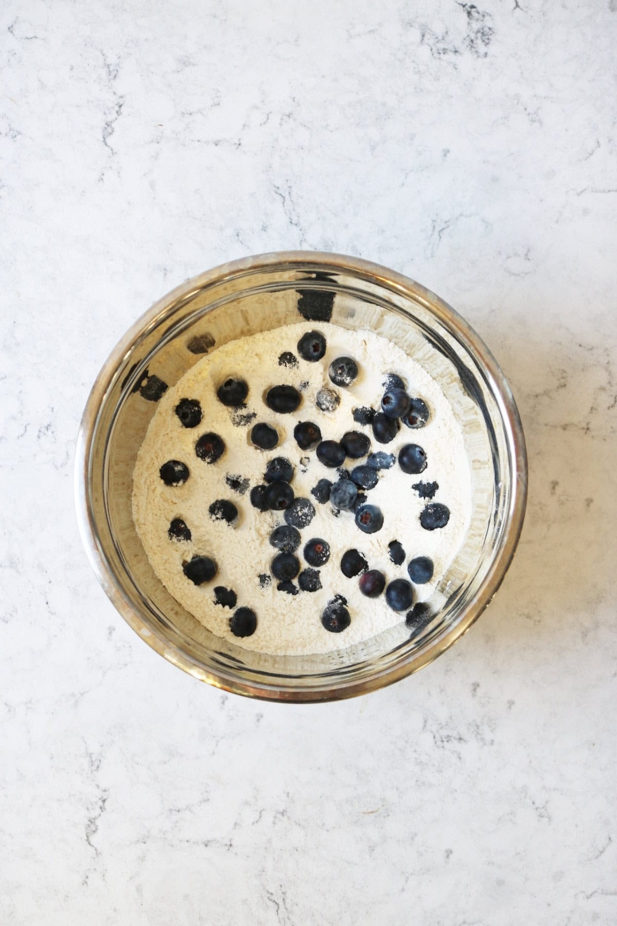 Flour, caster sugar and blueberries in a metal mixing bowl on a white marble counter top