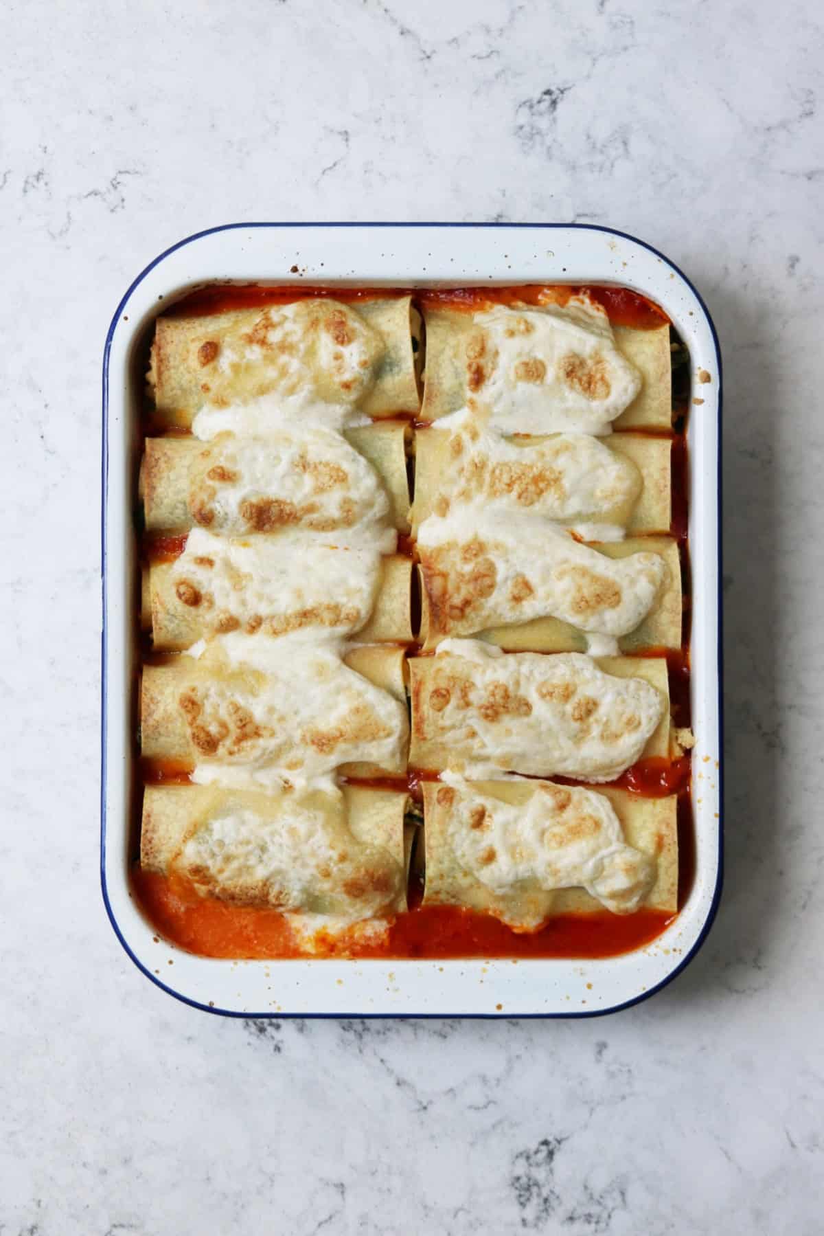 A blue and white enamel oven tin filled with tomato sauce and rolled up lasagne sheets stuffed with spinach and vegan ricotta, topped with mozzarella and baked until golden