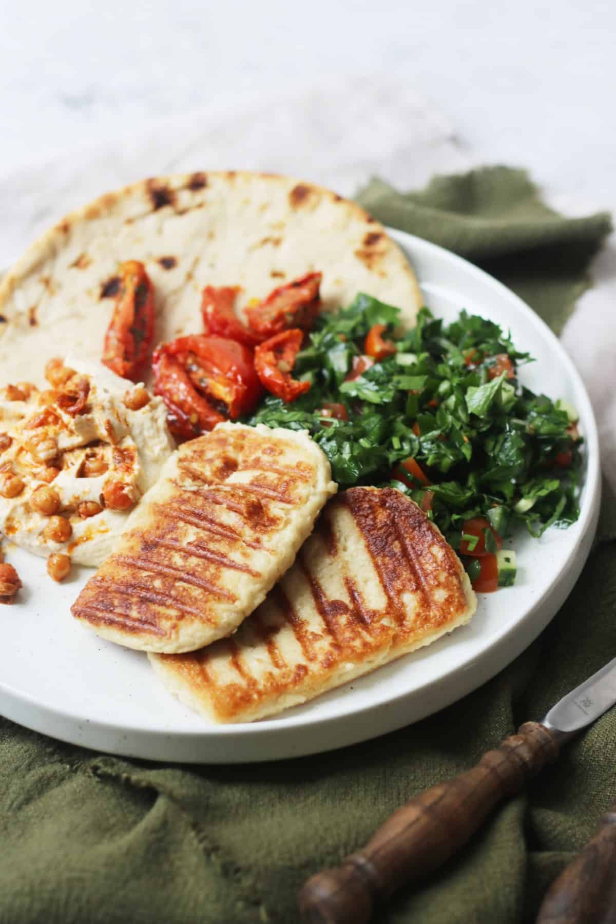 A plate topped with a flatbread, hummus and chickpeas, tabbouleh, sun dried tomatoes and vegan halloumi that has been grilled