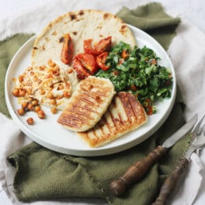 A plate topped with a flatbread, hummus and chickpeas, tabbouleh, sun dried tomatoes and vegan halloumi that has been grilled