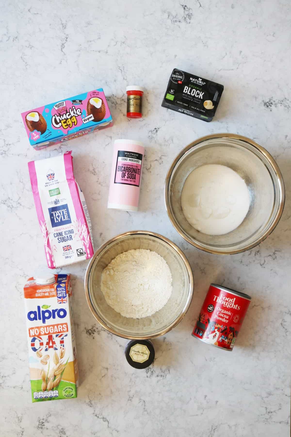 Vegan creme eggs, yellow food colouring, Naturli block vegan butter, icing sugar, bicarbonate of soda, caster sugar, oat milk, self raising flour, cocoa powder and vanilla paste on a white marble counter top