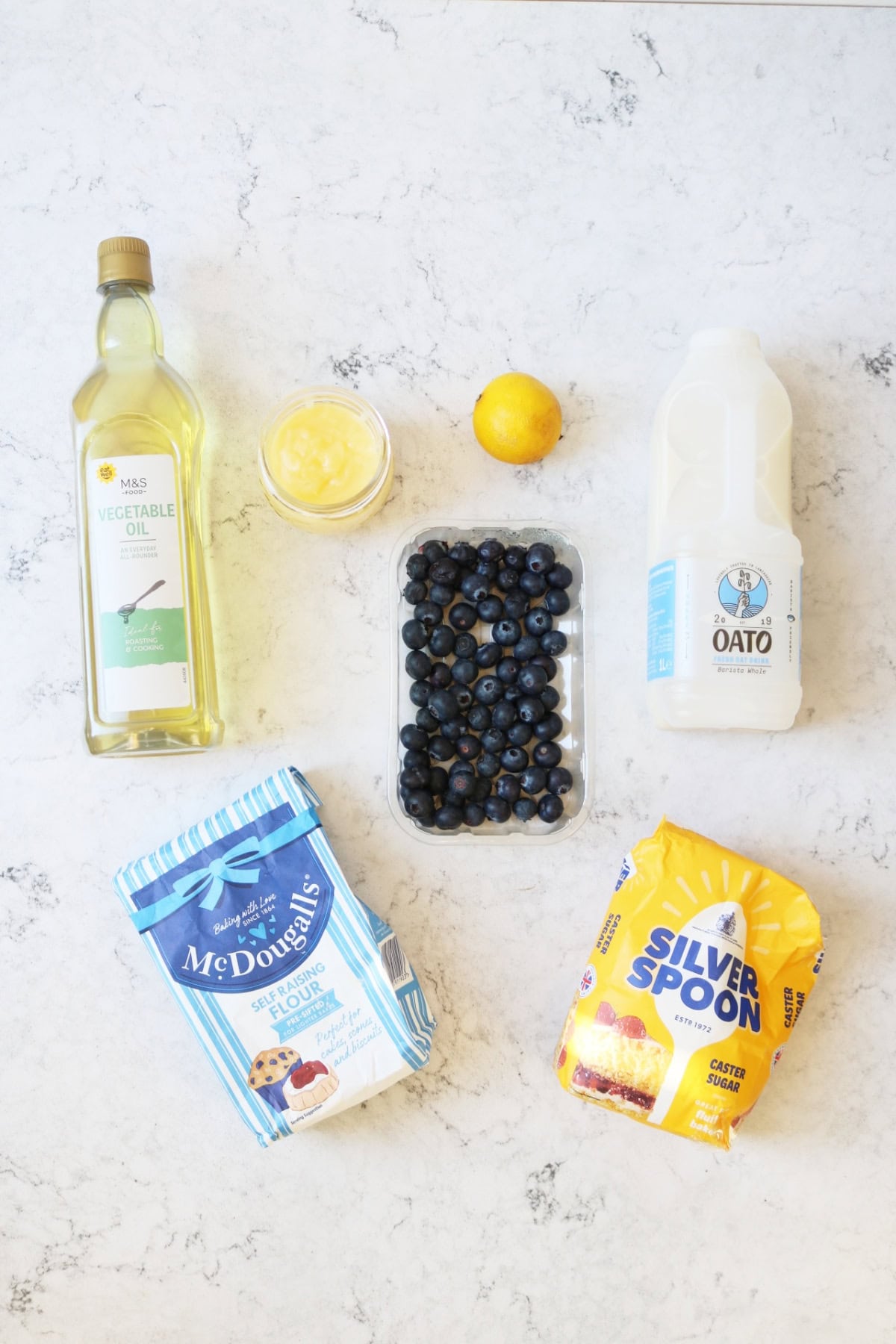 Vegetable oil, lemon curd, lemon, oat milk, blueberries, self raising flour and caster sugar laid out on a white marble counter top