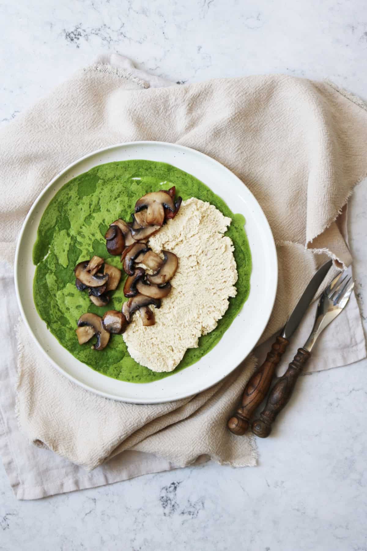 A spinach crêpe on a plate topped with vegan ricotta and cooked mushrooms
