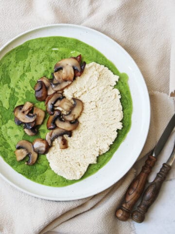 A spinach crêpe on a plate topped with vegan ricotta and cooked mushrooms and with a knife and fork