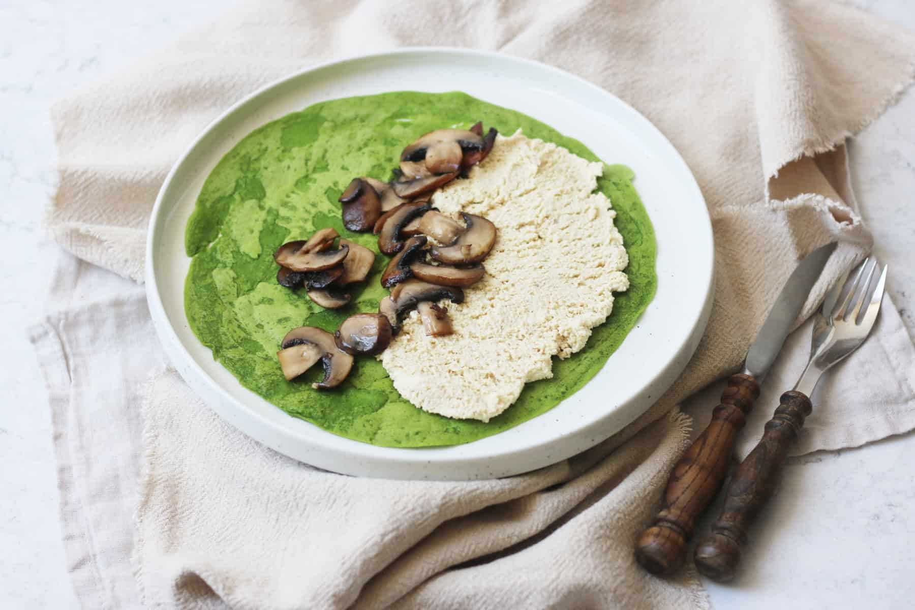 A spinach crêpe on a plate topped with vegan ricotta and cooked mushrooms