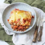 A square portion of vegan lentil lasagne on a blue plate that is sitting on top of green and cream napkins with a knife and fork.