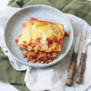 A square portion of vegan lentil lasagne on a blue plate that is sitting on top of green and cream napkins with a knife and fork.