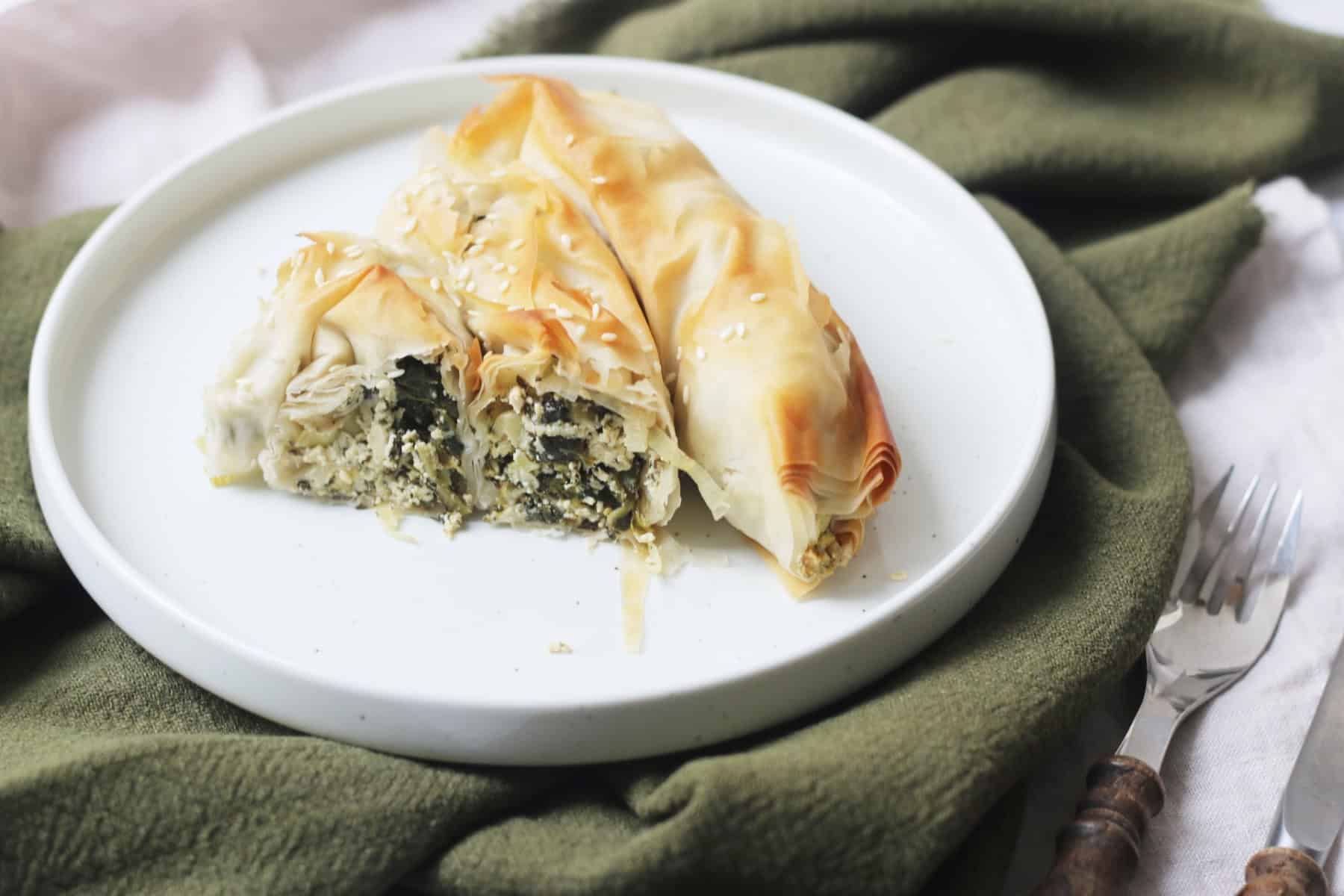A slice of Vegan Spiral Spanakopita on a white plate on top of a green napkin with a knife and fork to the right of the plate