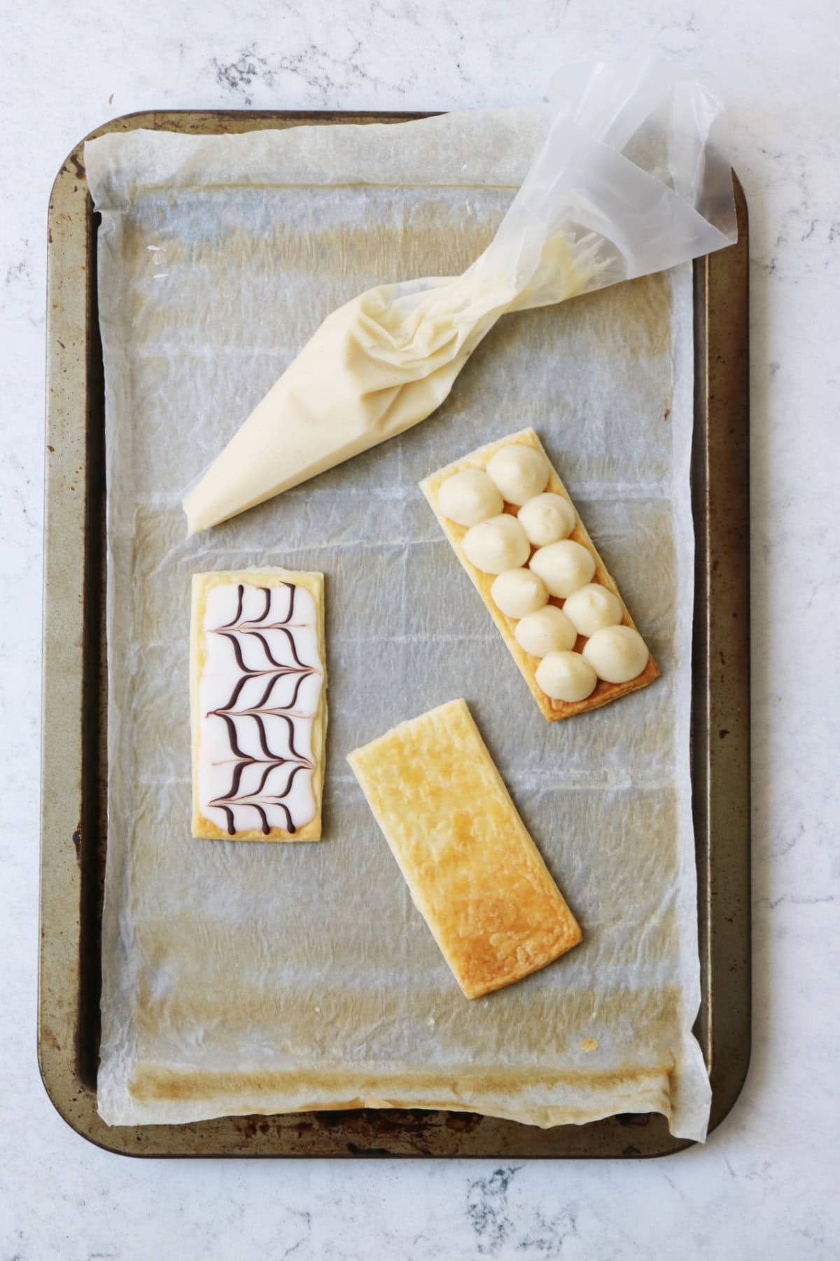 3 rectangles of puff pastry on a baking paper lined baking tray along with a piping bag full of vegan creme patissiere. One rectangle of puff pastry is topped with piped creme patissiere. A second rectangle of puff pastry is decorated with white icing and feathered chocolate.
