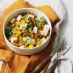 A bowl of tofu scramble with vegan feta on top of a yellow napkin on a white marble counter. A knife and fork are to the right of the bowl.
