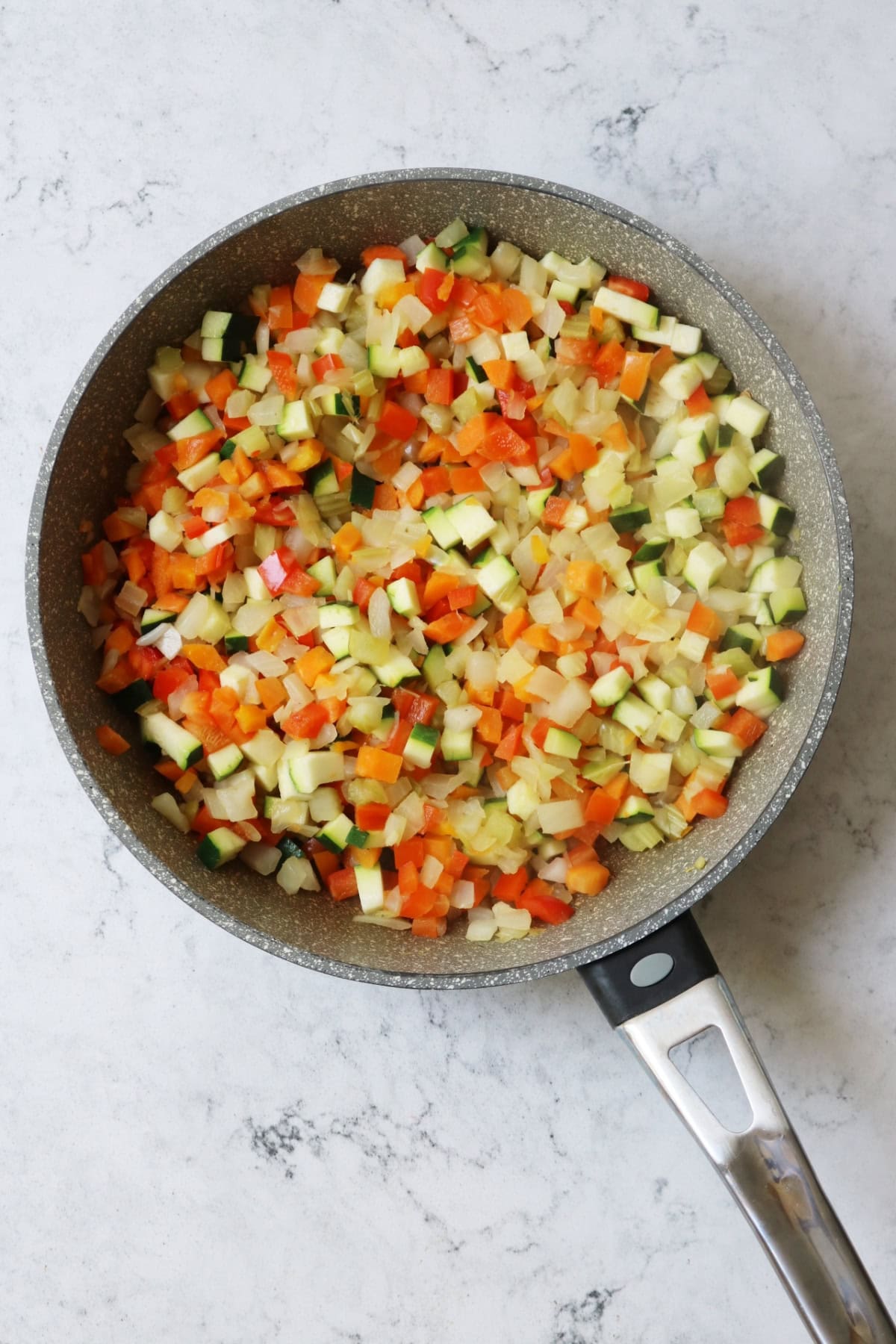 Diced onion, carrot, celery, red pepper and courgette in a frying pan on a white marble counter top.