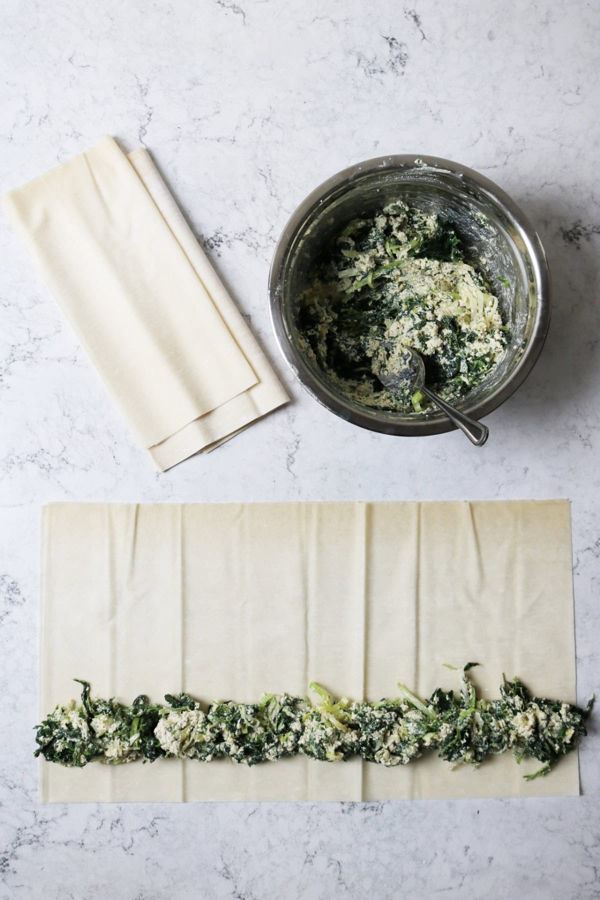 Sheets of filo pastry and a bowl of vegan spanakopita filling on a white marble counter top. Some of the filling has been spooned on to the filo pastry sheet running horizontally