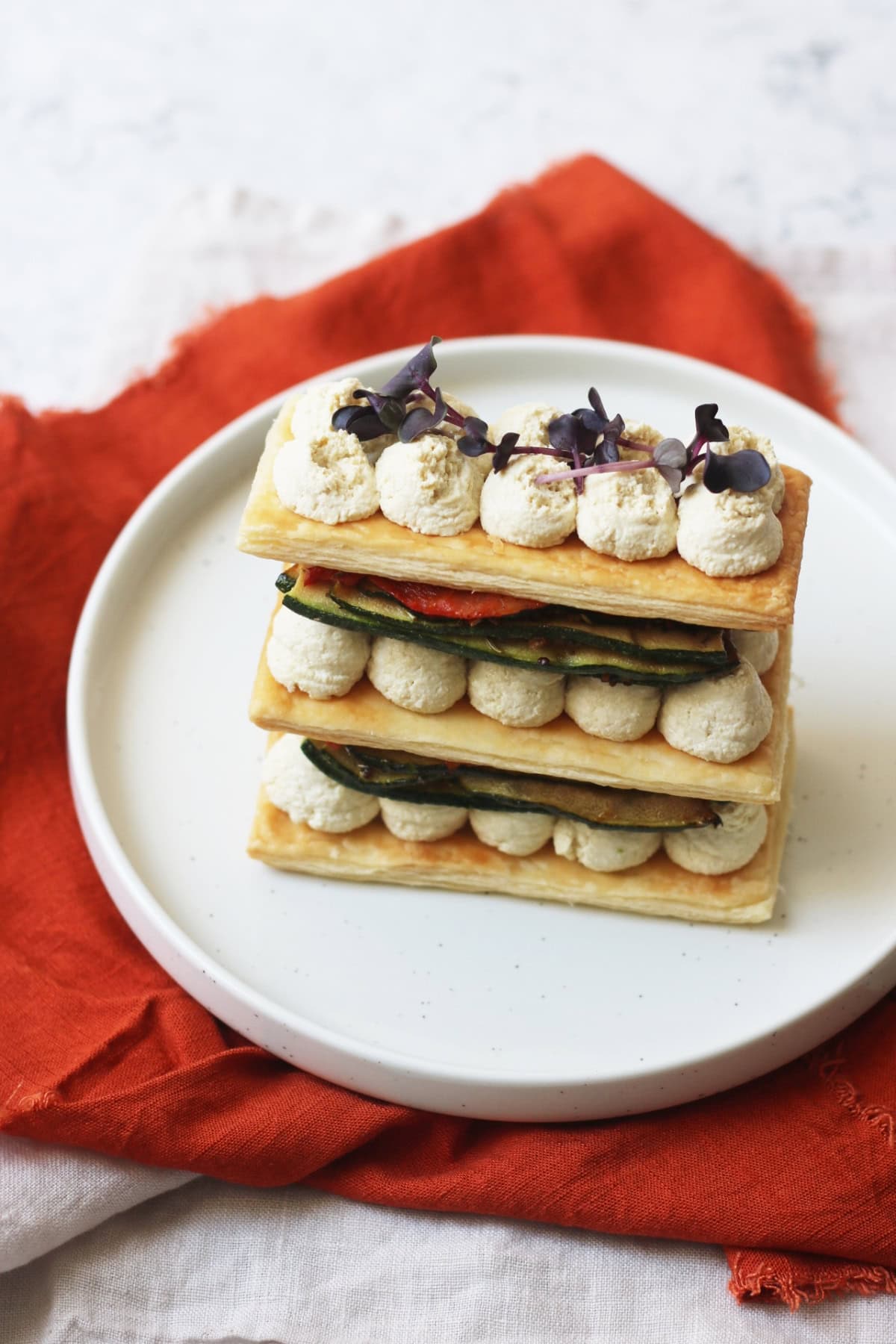 A Vegan Grilled Vegetable Mille-Feuille topped with micro herbs, on a plate with an orange napkin