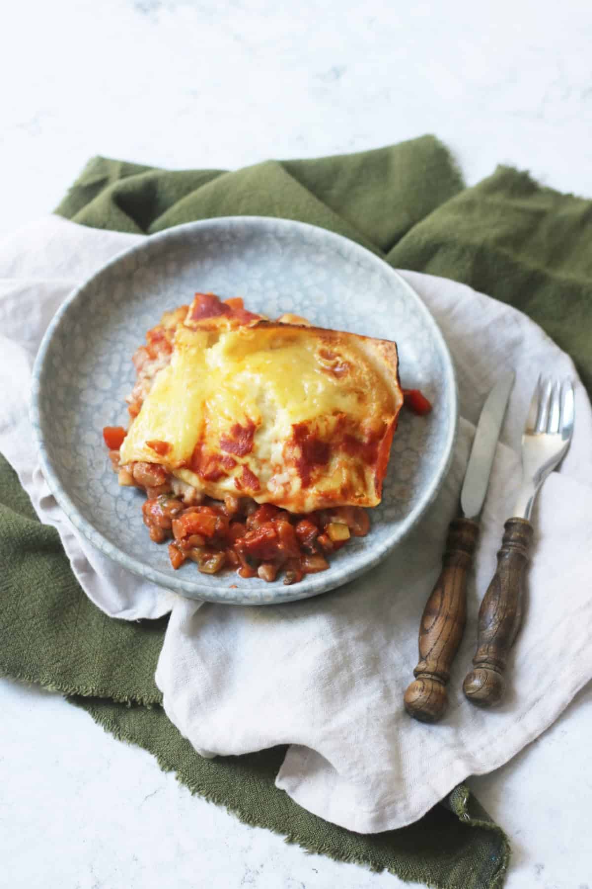 A square portion of vegan lentil lasagne on a blue plate that is sitting on top of green and cream napkins with a knife and fork.