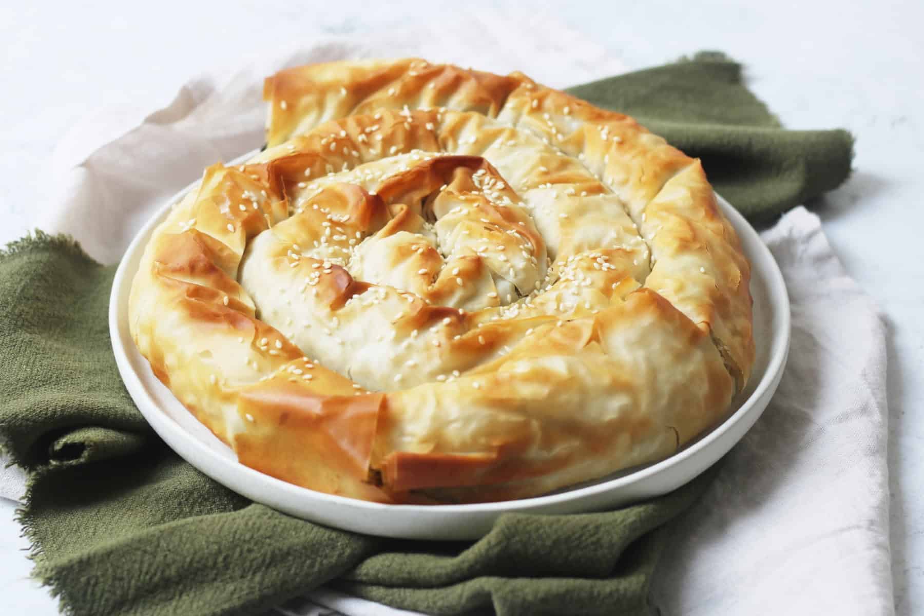 A baked Vegan Spiral Spanakopita on a plate on top of green and white napkins on a white marble counter top