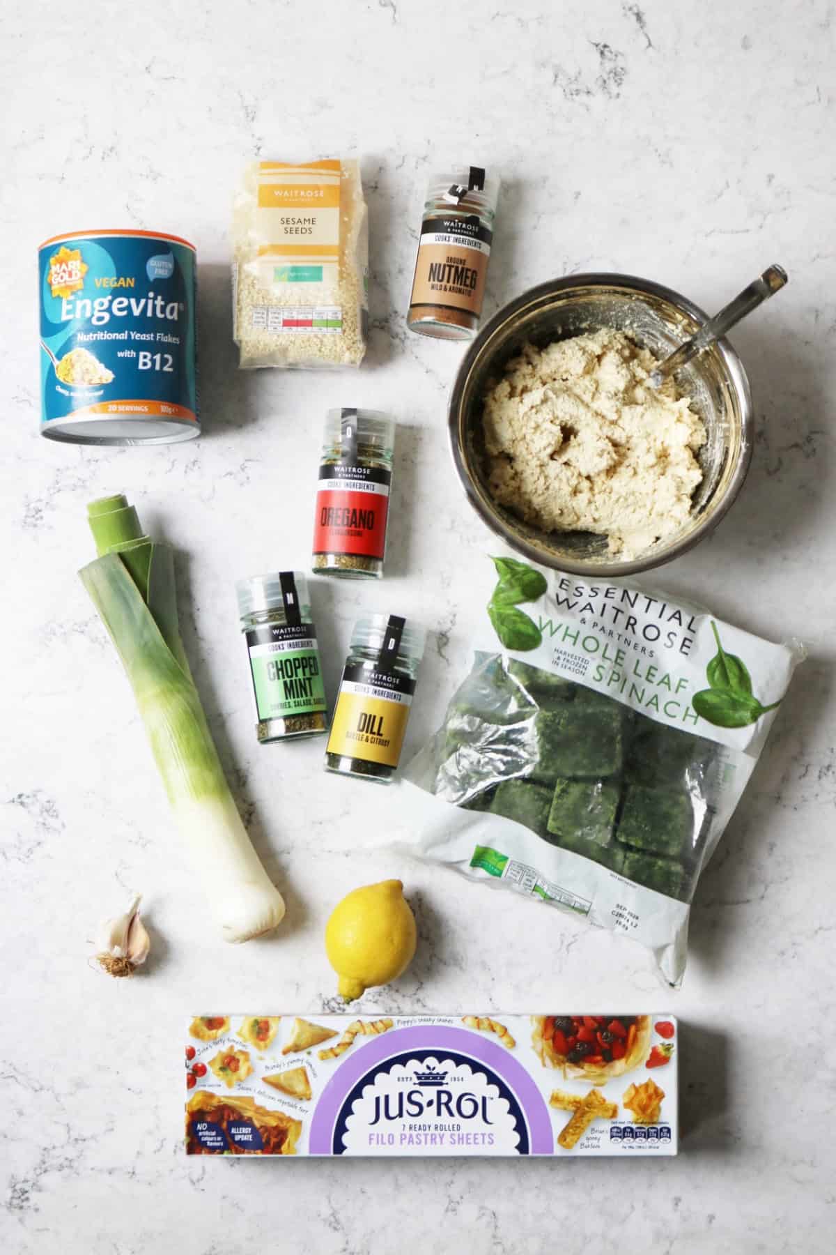Nutritional yeast, sesame seeds, nutmeg, vegan ricotta, oregano, leek, mint, dill, frozen spinach, garlic, lemon and filo pastry laid out on a white marble counter top.