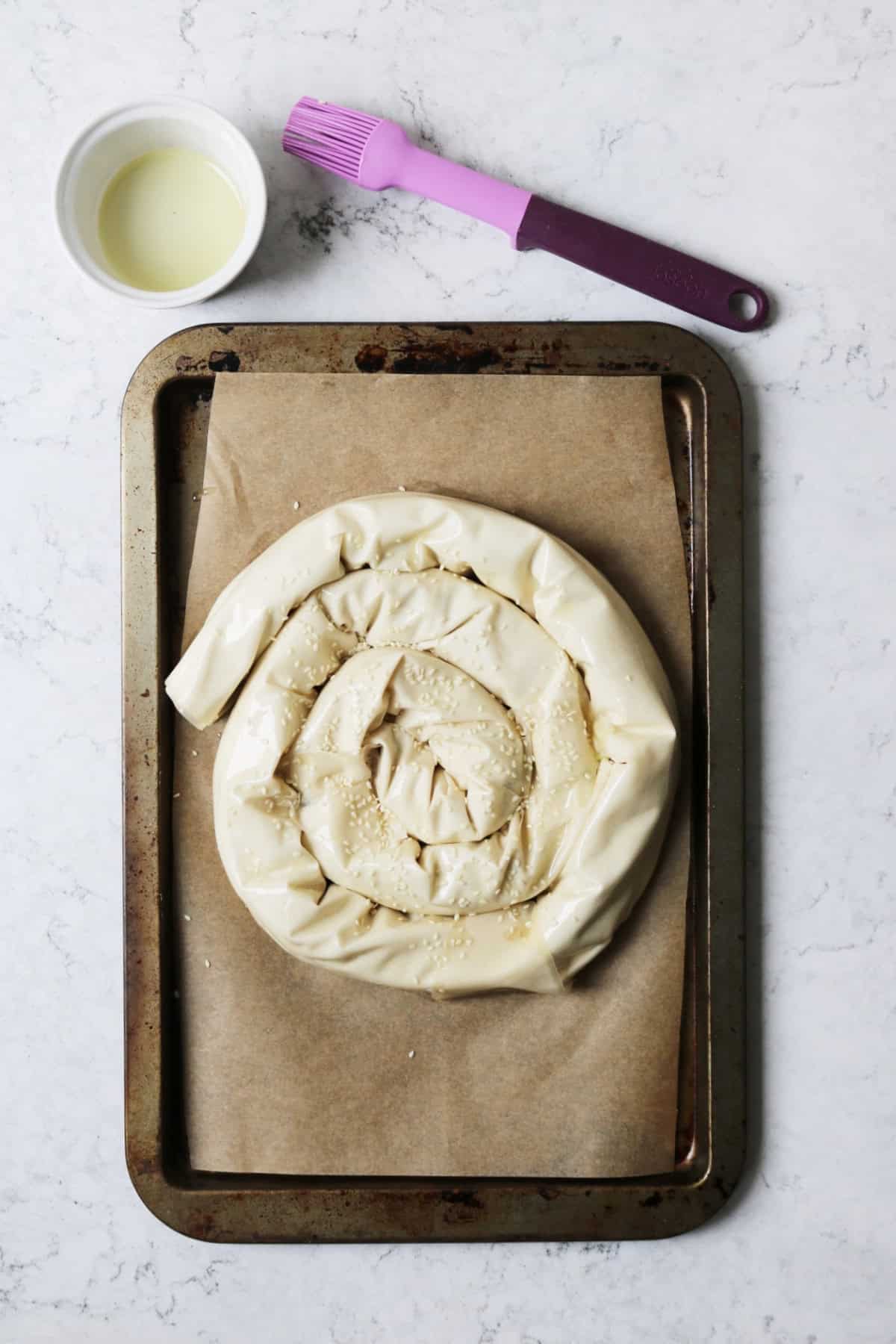 A spiral vegan spanakopita ready for the oven with a small pot of oil and a purple pastry brush
