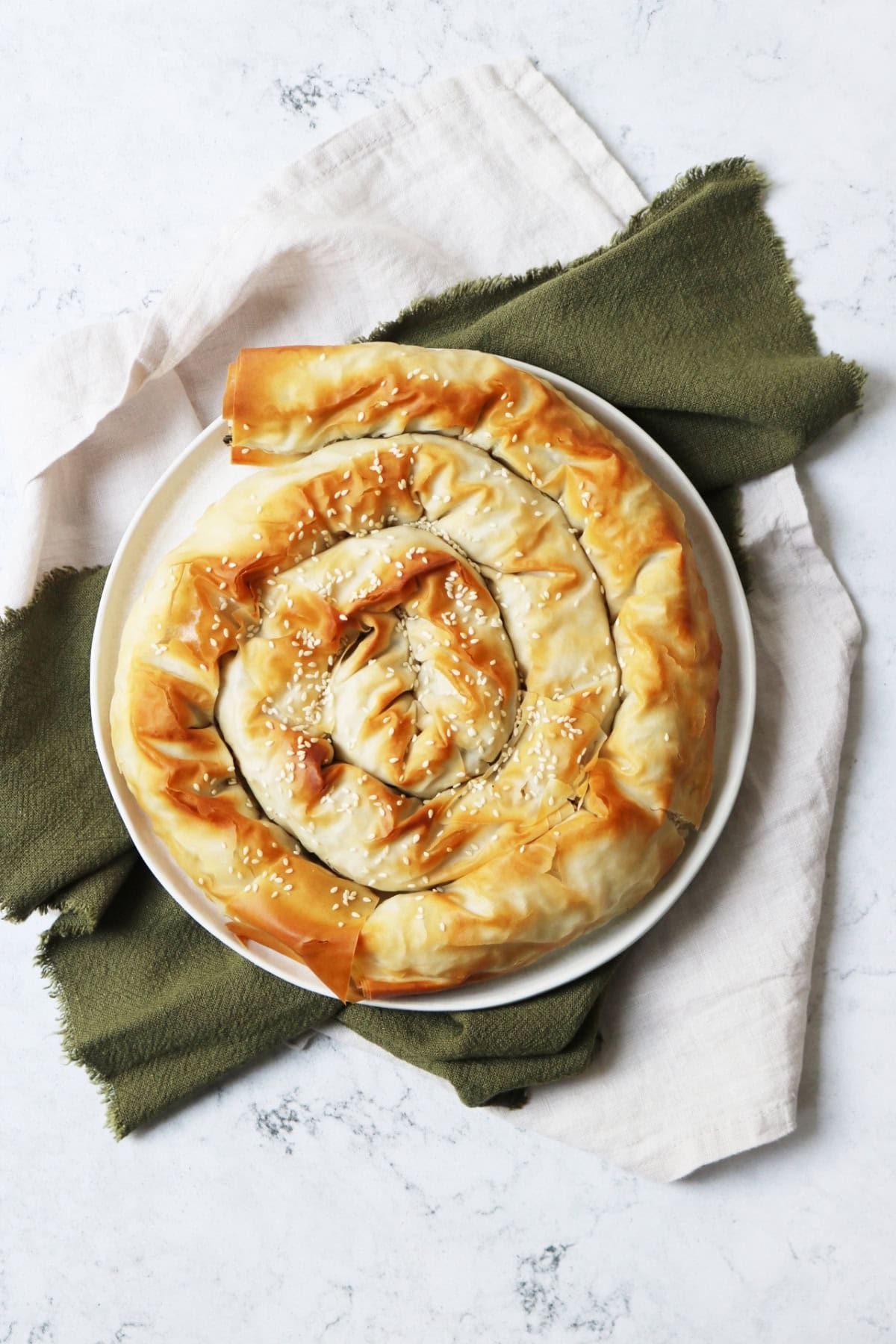 A baked Vegan Spiral Spanakopita on a plate on top of green and white napkins on a white marble counter top