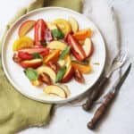 A tomato, peach and basil salad made up of wedges of heirloom tomatoes and fresh peach topped with basil leaves and balsamic vinegar on a large white plate