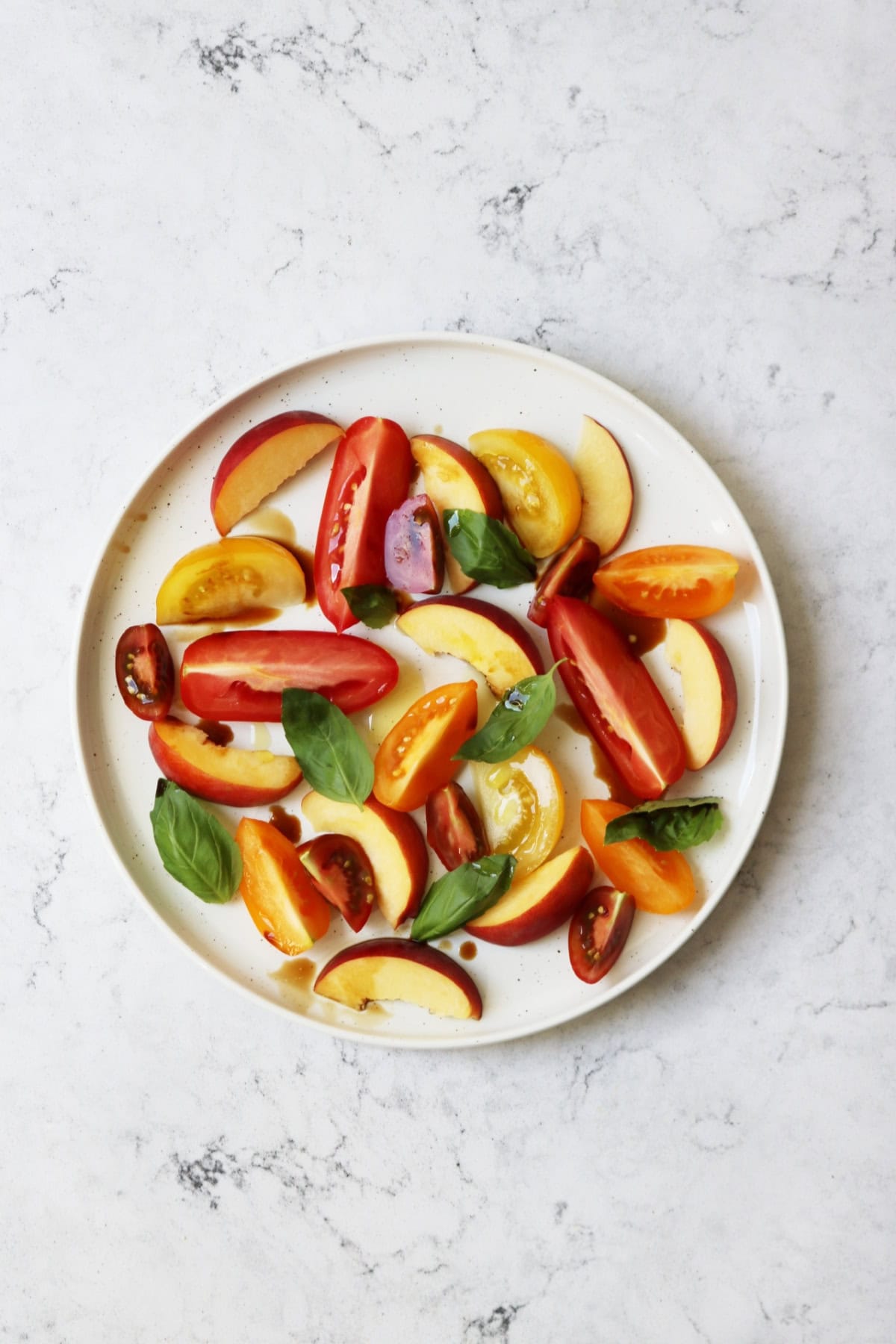 A tomato, peach and basil salad made up of wedges of heirloom tomatoes and fresh peach topped with basil leaves and balsamic vinegar on a large white plate