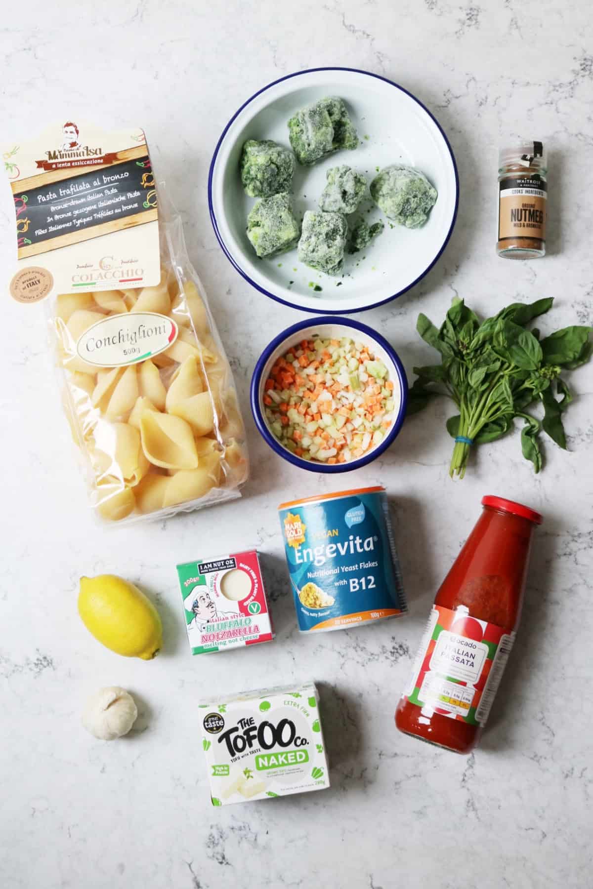 Jumbo pasta shells, frozen spinach, nutmeg, diced carrot, celery and onion, fresh basil, lemon, vegan mozzarella, nutritional yeast, garlic, tofu, passata laid out on a white marble counter top