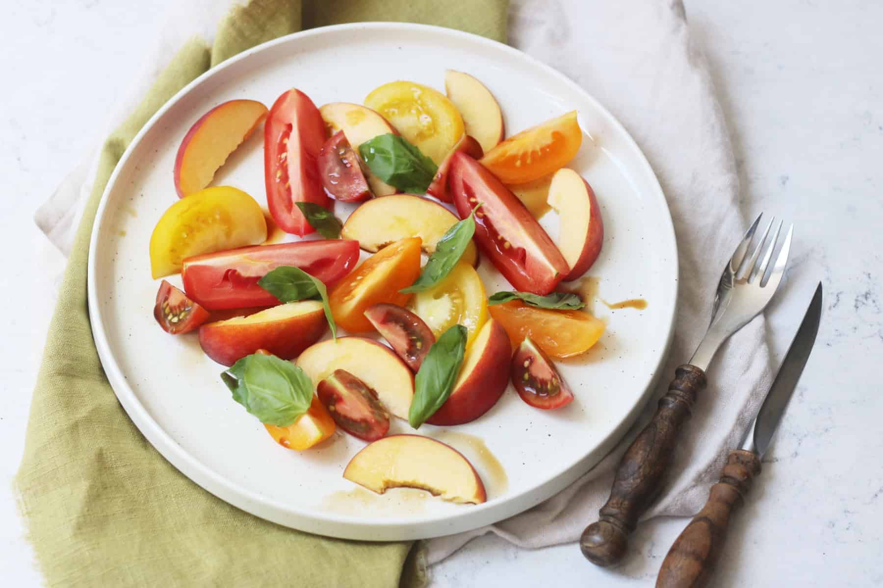 A tomato, peach and basil salad made up of wedges of heirloom tomatoes and fresh peach topped with basil leaves and balsamic vinegar on a large white plate