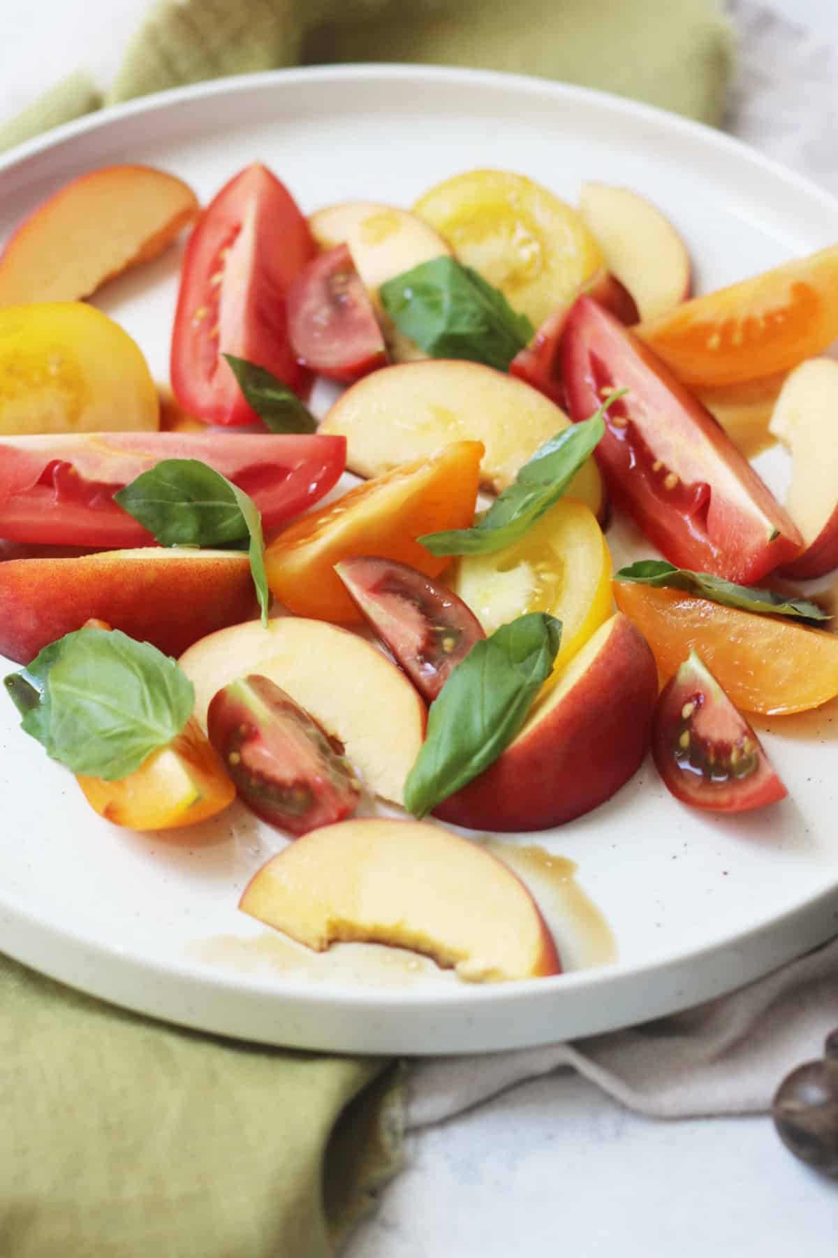 A tomato, peach and basil salad made up of wedges of heirloom tomatoes and fresh peach topped with basil leaves and balsamic vinegar on a large white plate