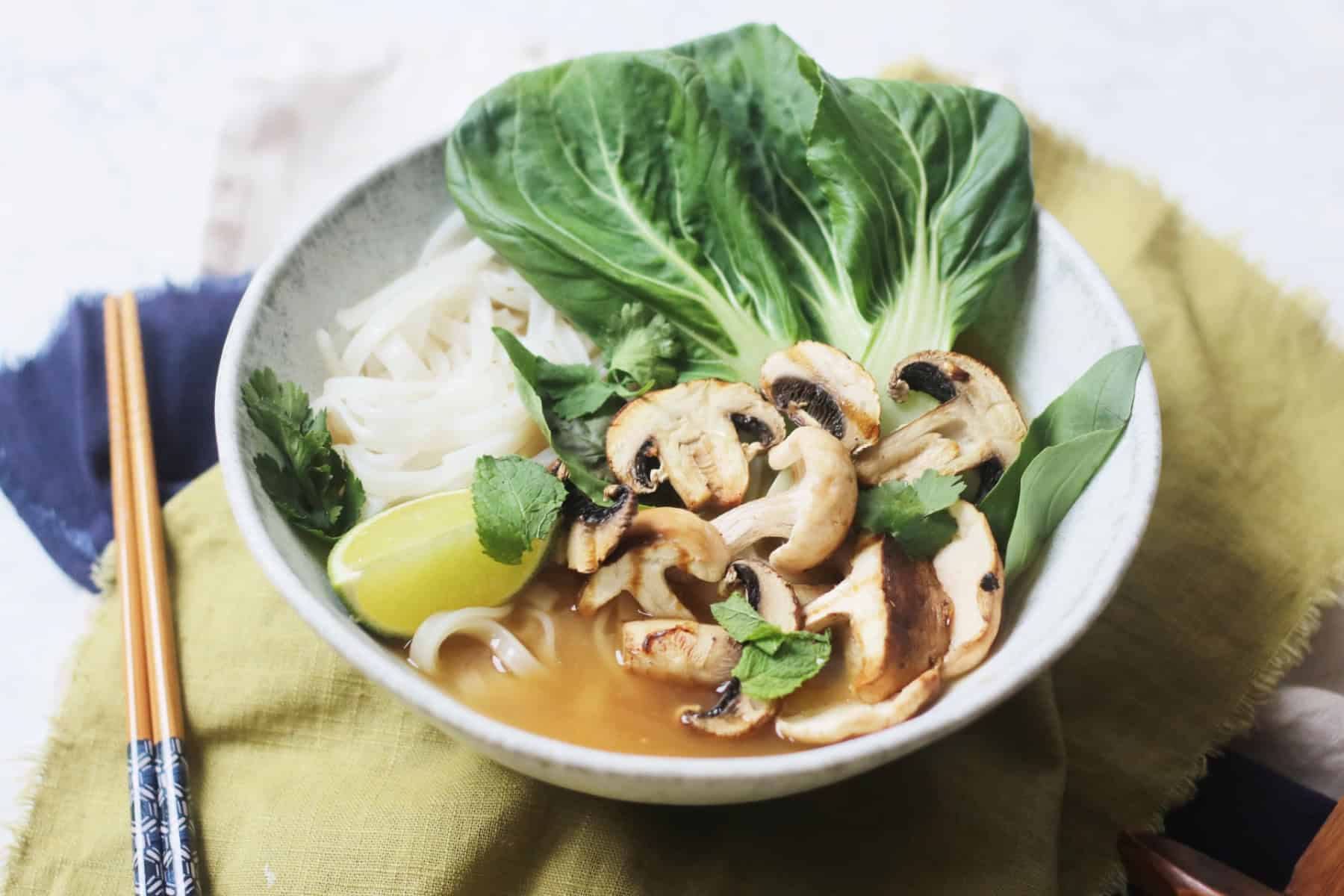A bowl of Vegan Pho topped with pak choi, mushrooms, fresh herbs and a wedge of lime