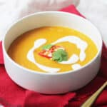 Vegan Carrot and Ginger Soup in a white bowl, served with a drizzle of yogurt, red chilli and coriander