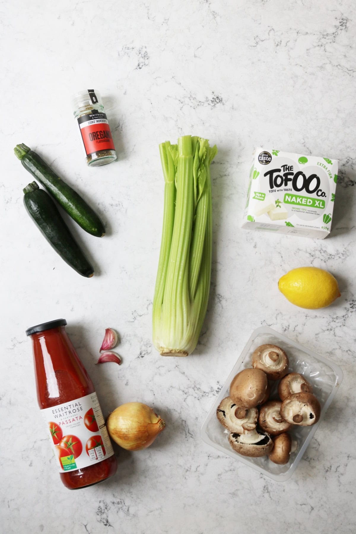 Dried oregano, courgette, celery, tofu, lemon, garlic, passata, onion, mushrooms laid out on a white marble counter top